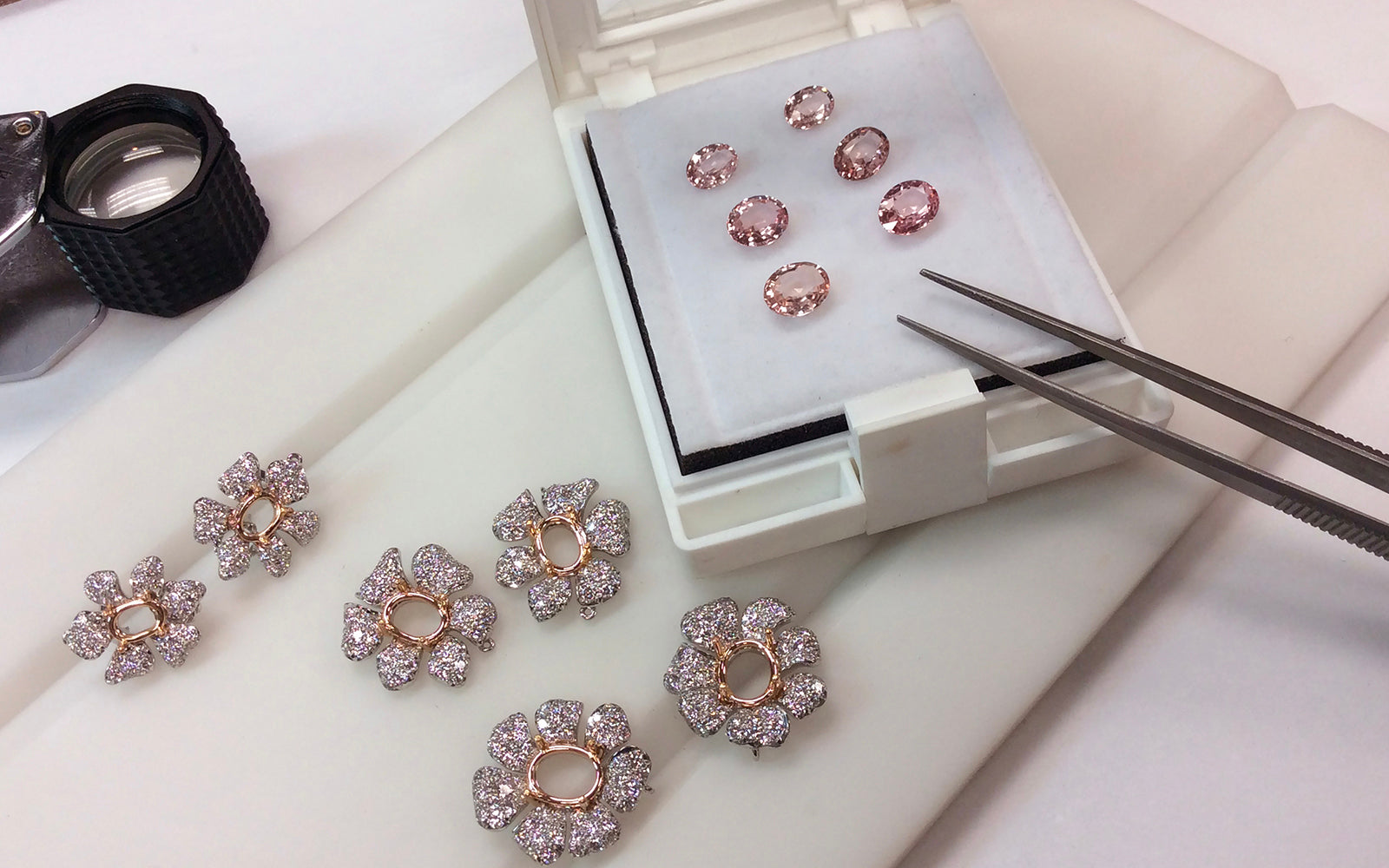Diamond in pink gold flower settings and pink gems in a box with tweezers and a jewelry loupe