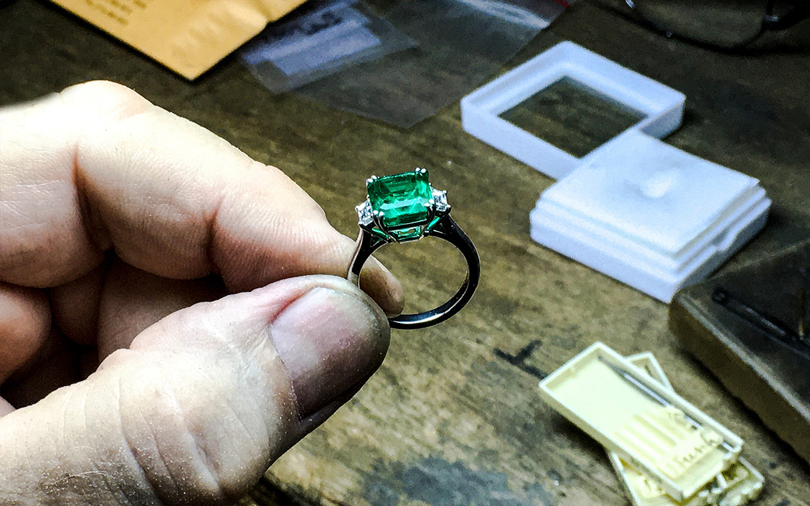 Hand holding emerald and diamond ring over wooden workbench