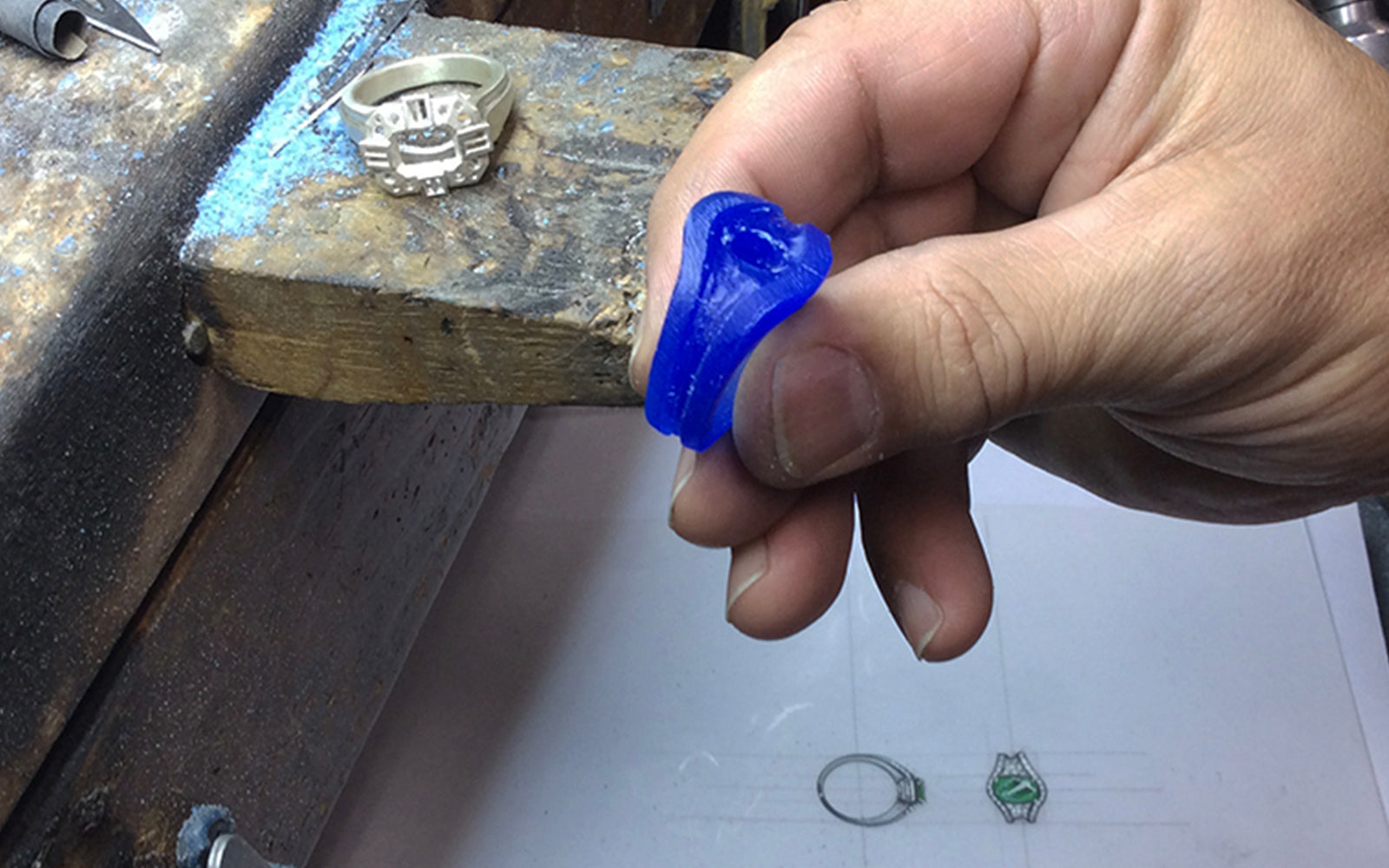 Jeweler's hand holding wax carving over workbench with unfinished ring and sketch