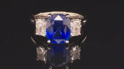 Video of rotating platinum ring with a blue sapphire and two radiant cut diamonds on either side on black background.