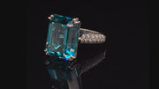 Video of rotating platinum ring with emerald-cut blue zircon and round diamonds on black background.