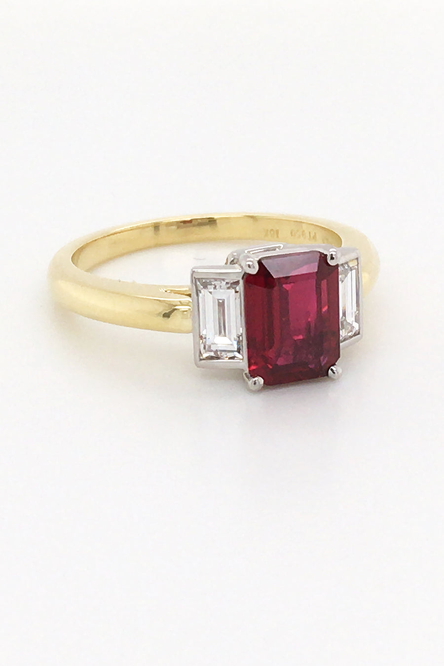 Angle view of three-stone ruby ring with two baguette diamonds set in platinum on a yellow gold band on a white background