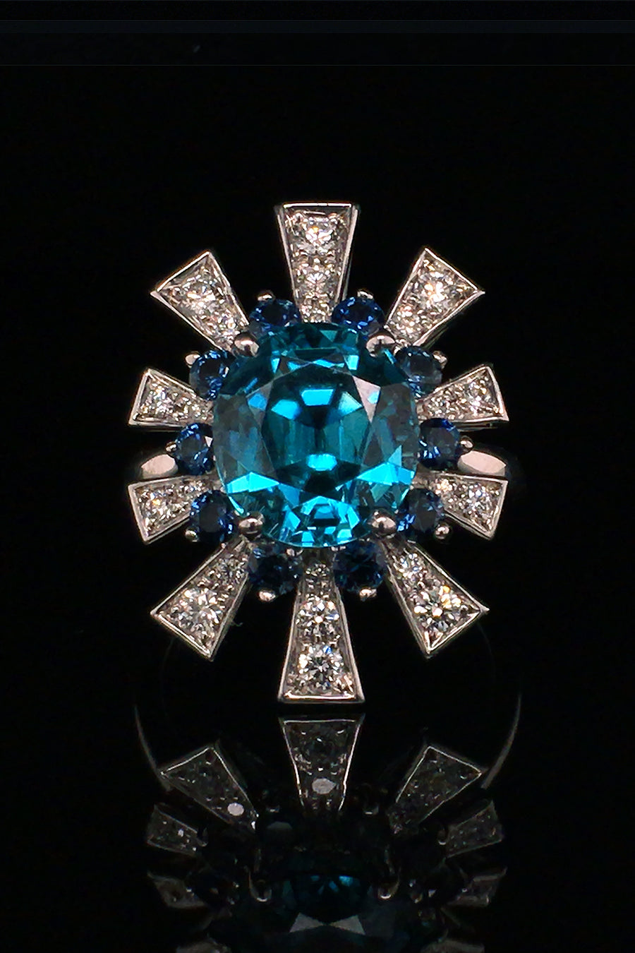 18K white gold ring featuring a blue zircon surrounded by a starburst motif of blue sapphires and brilliant diamonds on a black background.