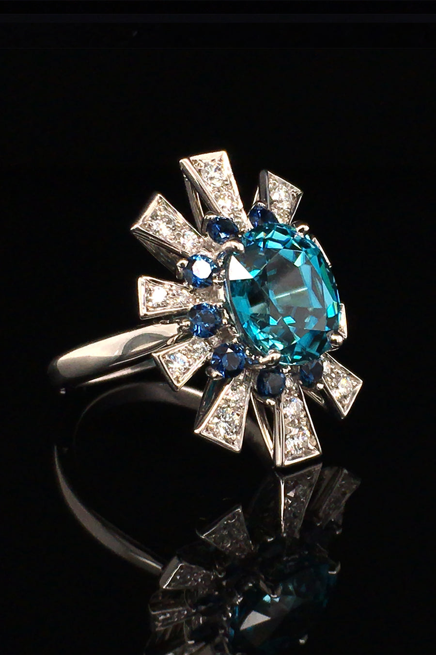 Angle view of 18K white gold ring featuring a blue zircon surrounded by a starburst motif of blue sapphires and brilliant diamonds on a black background.