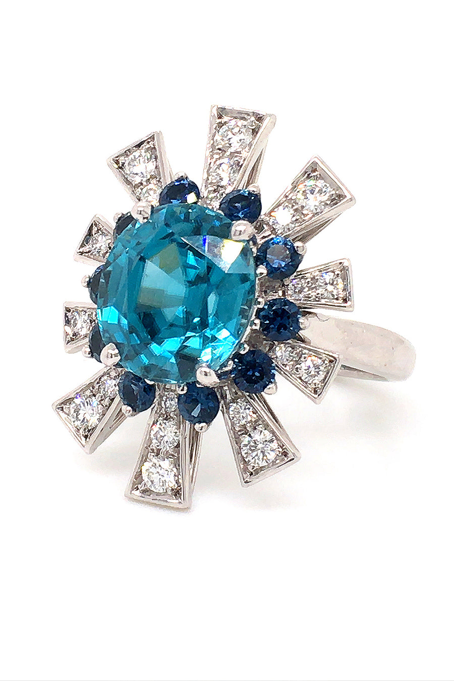 Angle view of 18K white gold ring featuring a blue zircon surrounded by a starburst motif of blue sapphires and brilliant diamonds on a white background.