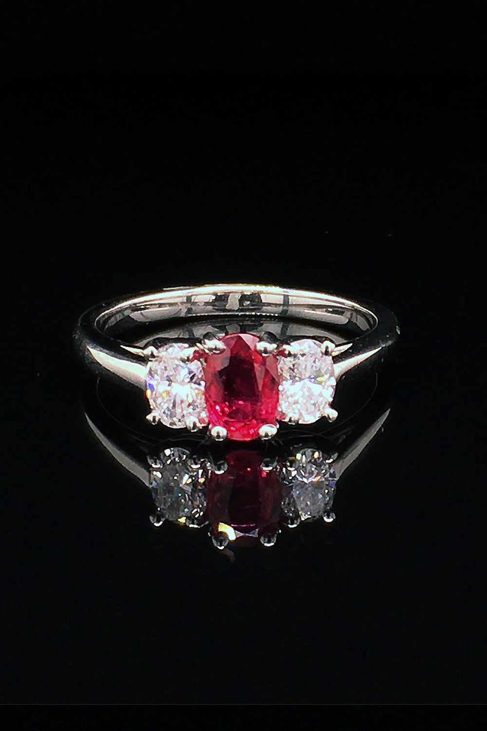 Platinum three-stone ring with a oval ruby flanked by two oval diamonds on a black background.