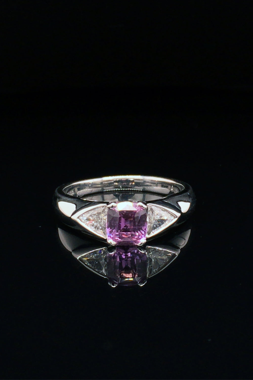 Platinum ring with pink sapphire and two trilliant diamonds on black background.