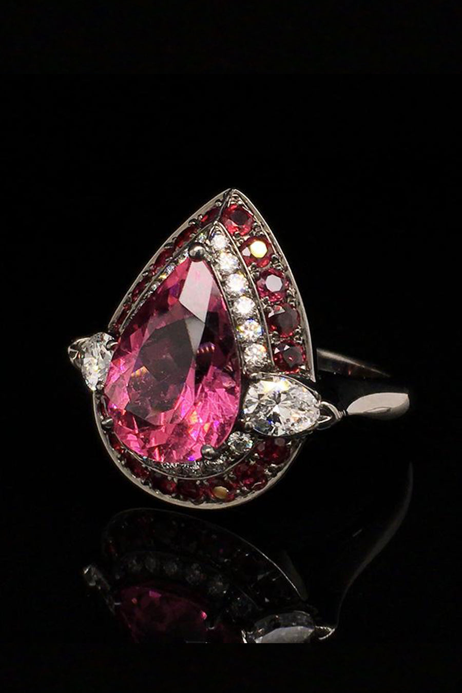 Angle view of 18K white gold ring with a pear-shape rubellite surrounded by diamonds and rubies with a black rhodium finish on a black background