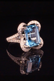 Angle view of 18K white gold ring with an emerald-cut aquamarine and diamonds in a ribbon motif on a black background