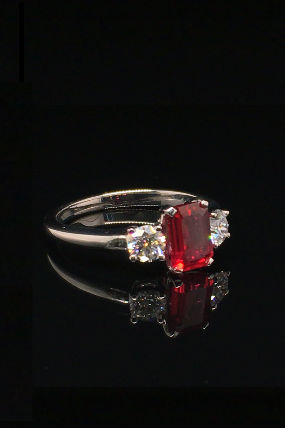 Angle view of platinum ring with an emerald-cut ruby and two round diamonds on a black background.