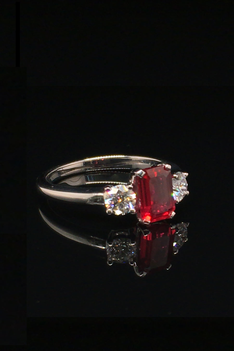 Angle view of platinum ring with an emerald-cut ruby and two round diamonds on a black background.