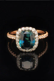 20K pink gold ring with a cushion-cut teal sapphire and diamond halo on a black background.