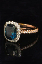 Angle view of 20K pink gold ring with a cushion-cut teal sapphire and diamond halo on a black background.