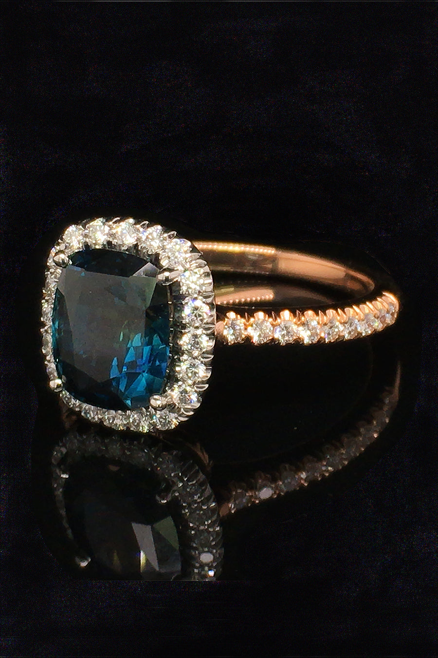 Angle view of 20K pink gold ring with a cushion-cut teal sapphire and diamond halo on a black background.