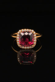20K pink gold ring with a cushion-shaped rhodolite garnet and diamond halo on a black background.
