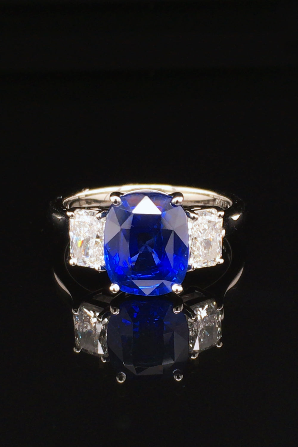 Platinum ring with a blue sapphire and two radiant cut diamonds on either side on black background.