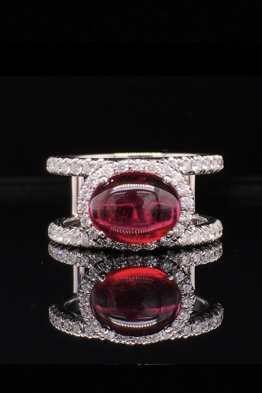18K white gold ring with an oval rubellite cabochon in a diamond halo between two diamond bands on a black background