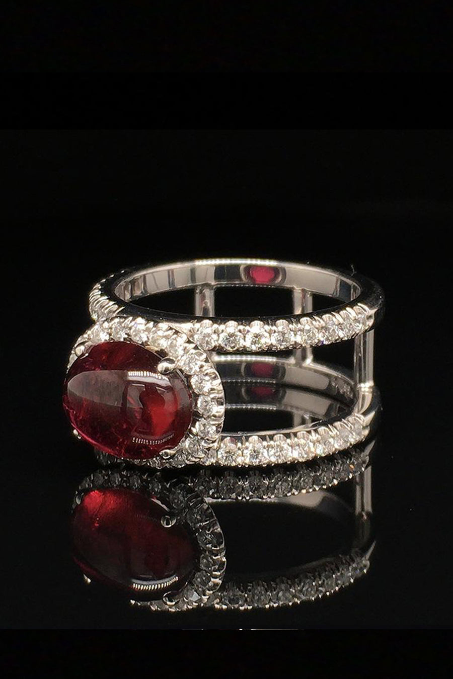 Angle view of 18K white gold ring with an oval rubellite cabochon in a diamond halo between two diamond bands on a black background