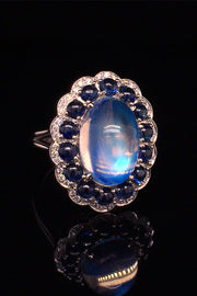 Angle view of platinum ring with a large moonstone surrounded by cabochon sapphires and diamonds on a black background