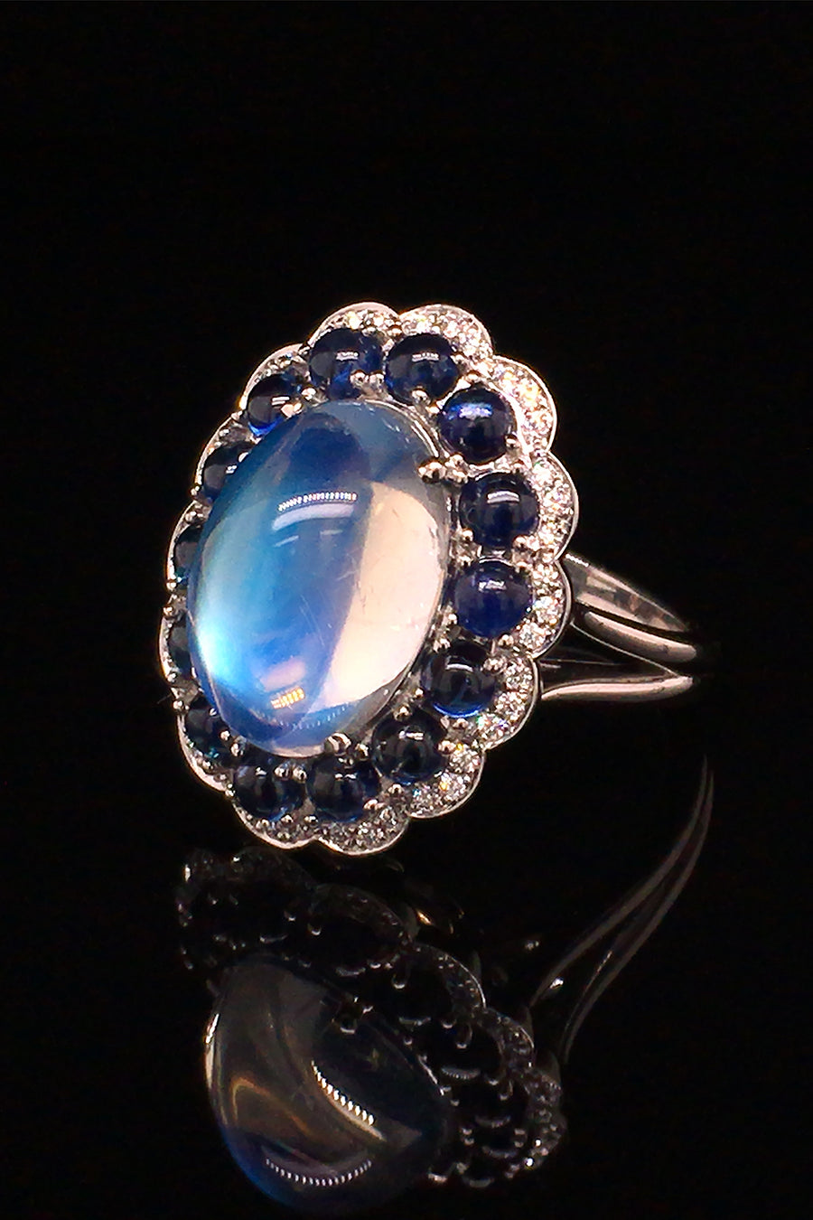 Angle view of platinum ring with a large moonstone surrounded by cabochon sapphires and diamonds on a black background