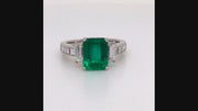 Video of rotating platinum ring with an emerald, baguette diamonds, and square diamonds on a white background