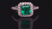 Video of rotating platinum ring with emerald and diamond halo on black background.