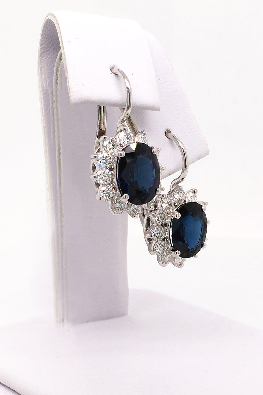 Angle view of 18K white gold earrings with oval blue sapphires surrounded by diamonds on white background