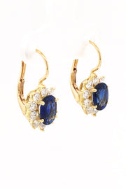 Angle of 18K yellow gold earrings featuring oval blue sapphires surrounded by smaller diamonds with lever back closures on white background