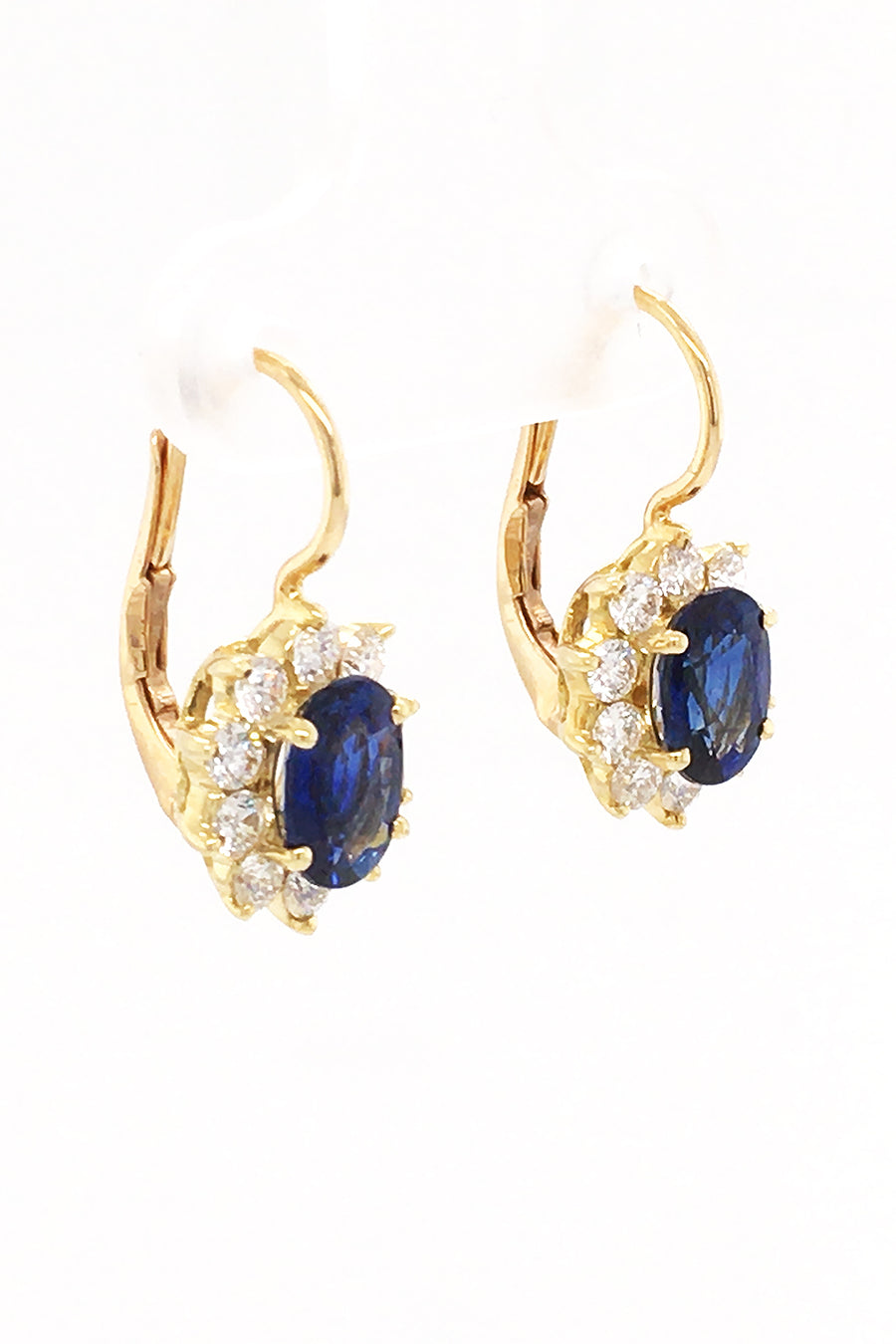 Angle of 18K yellow gold earrings featuring oval blue sapphires surrounded by smaller diamonds with lever back closures on white background