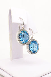 Angle view of 18K white gold earrings featuring large oval aquamarines surrounded with diamonds with lever backs on a white jewelry display