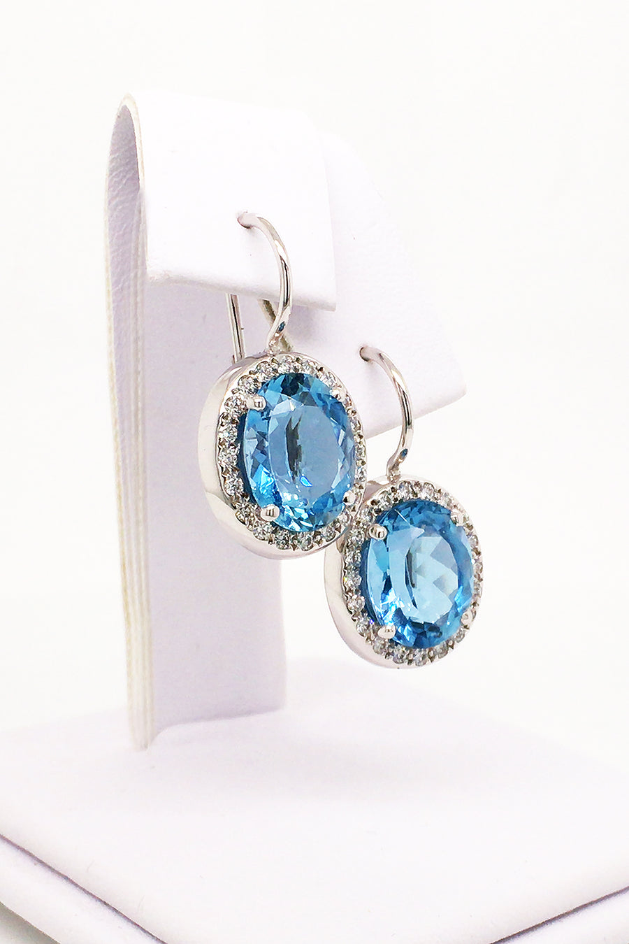 Angle view of 18K white gold earrings featuring large oval aquamarines surrounded with diamonds with lever backs on a white jewelry display