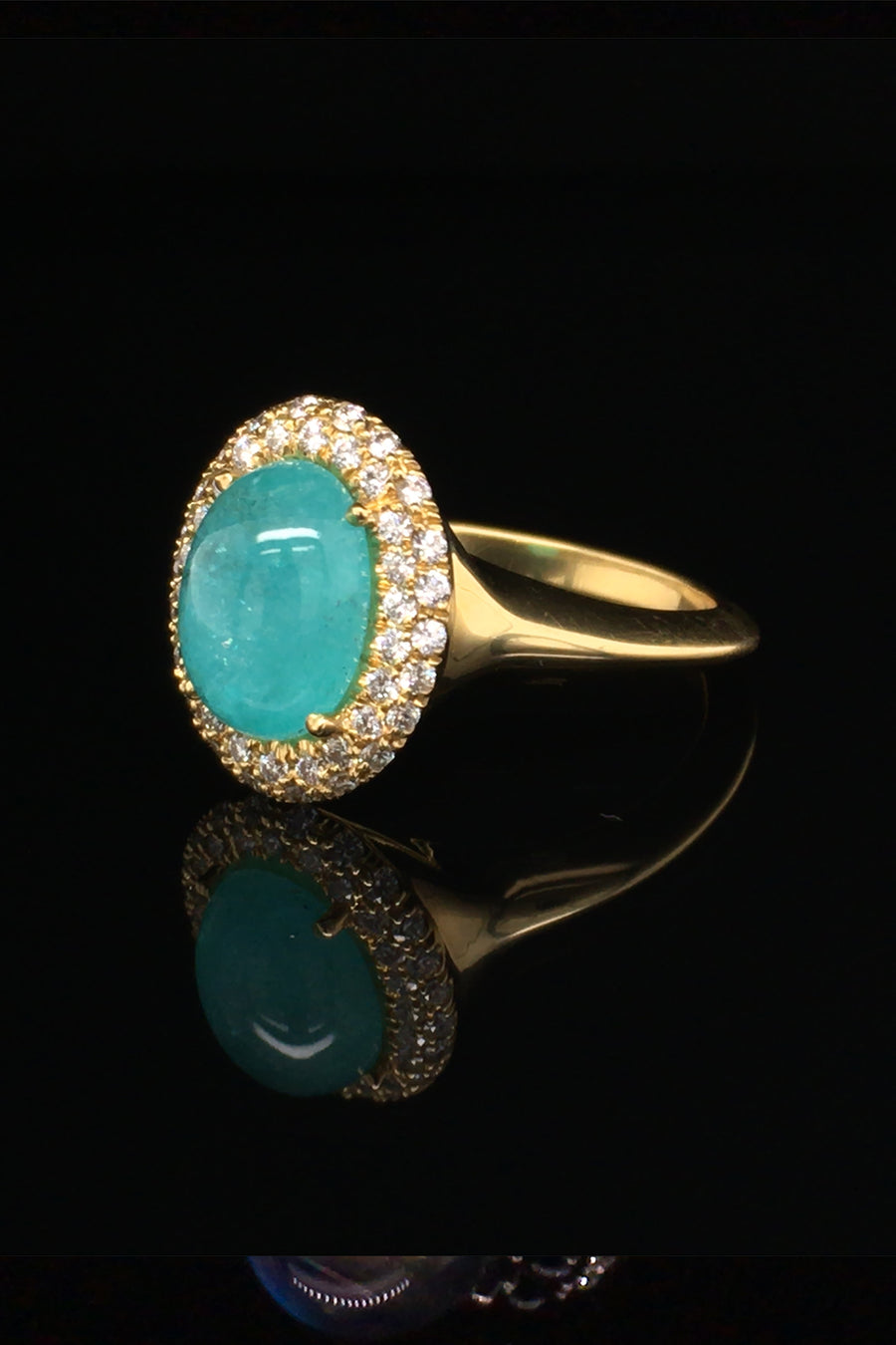 Angle view of 18K yellow gold ring with oval Brazilian paraiba cabochon and pave diamond halo on a black background.