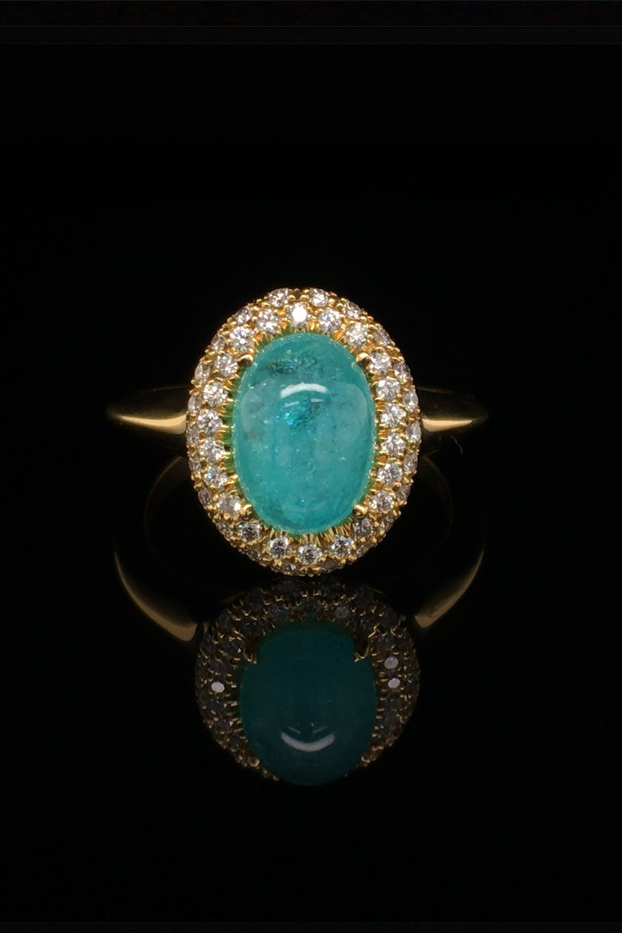 18K yellow gold ring with oval Brazilian paraiba cabochon and pave diamond halo on a black background.