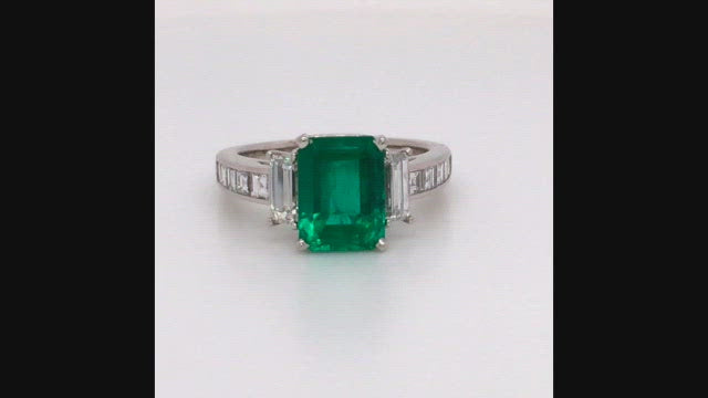 Video of rotating platinum ring with an emerald, baguette diamonds, and square diamonds on a white background